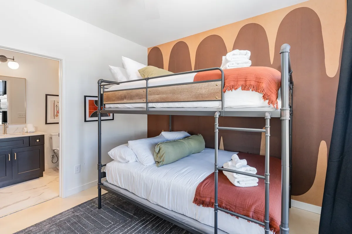 A cheerful bunk room featuring a bunk bed with two queen beds, colorful wall art, and a bathroom entrance in view. The playful yet comfortable setup invites group fun, game nights, and cozy sleepovers, appealing to both kids and adults.