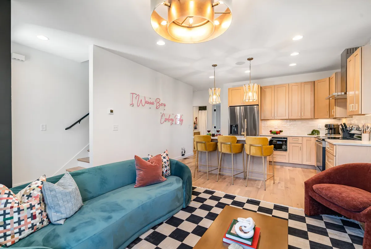 A bright and cheerful open-plan space showcasing a colorful sofa, a spacious kitchen island, and decorative wall art. The layout is designed for easy socializing, cooking, and entertaining.