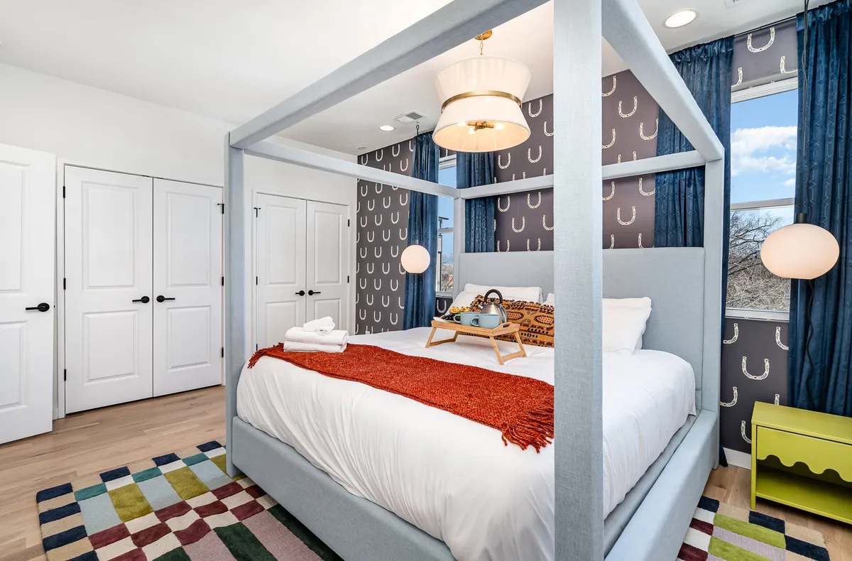 A cozy, inviting bedroom featuring a canopy bed with decorative pillows, a colorful rug, and a modern light fixture. The space feels bright and relaxing, ideal for morning coffee or a restful night after a day out. Storage options are visible, and the room is bathed in natural light from nearby windows.