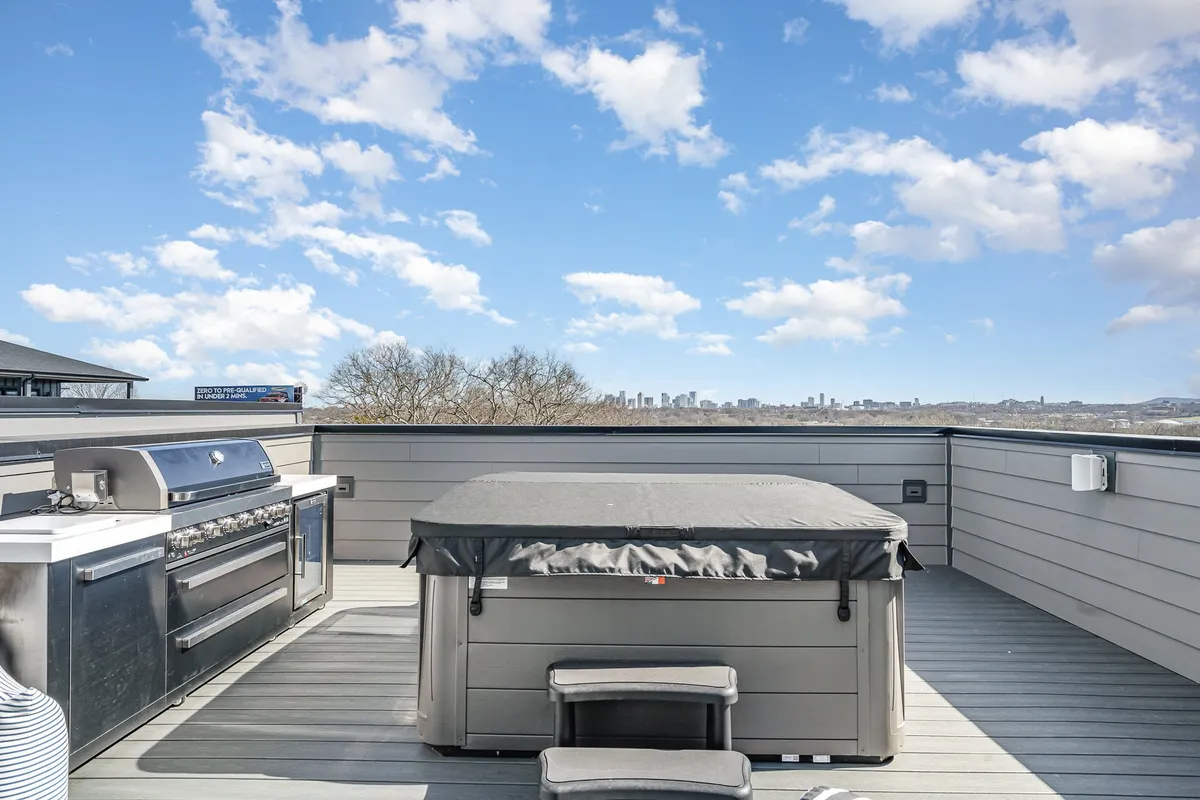 A modern rooftop deck featuring a bubbling hot tub, an outdoor kitchen with a grill, and comfortable seating with a stunning view of the Nashville city skyline in the background. The ambiance is inviting and perfect for socializing or unwinding in the hot tub while taking in the city lights.