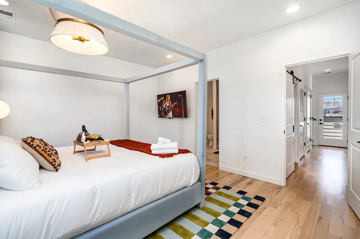 A stylish bedroom with a four-poster bed, vibrant rug, and colorful wall art. The space exudes both comfort and creativity, encouraging relaxation or quiet moments with a book. The overall mood is cozy and inviting, with tasteful decor.