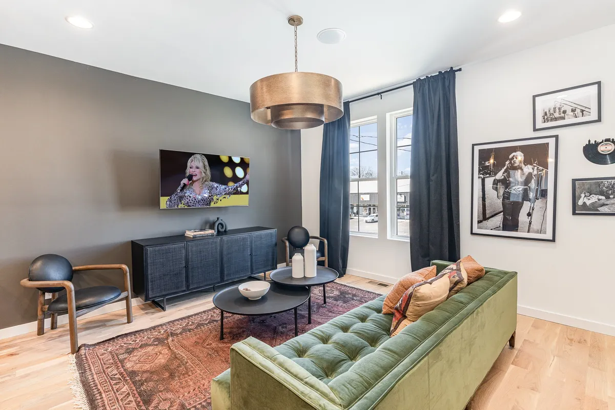 The image showcases a stylish and inviting living room featuring a plush green tufted sofa, a sleek black TV unit, vibrant artwork adorning the walls, and a modern hanging light fixture. The overall mood is contemporary yet comfortable, suggesting a perfect space for relaxation or gathering with loved ones.