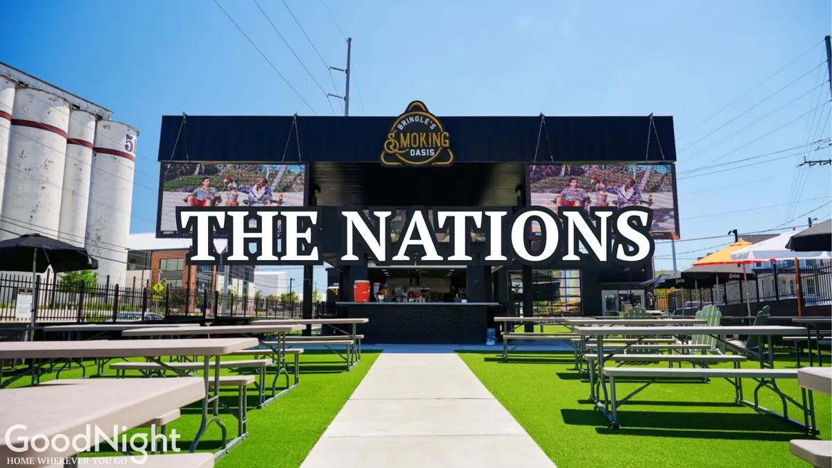 A festive street scene in The Nations neighborhood, this image shows a large sign reading 'THE NATIONS,' an outdoor seating area with colorful umbrellas, and a smoky grill area. The environment is lively, with people gathering for dining and community events.